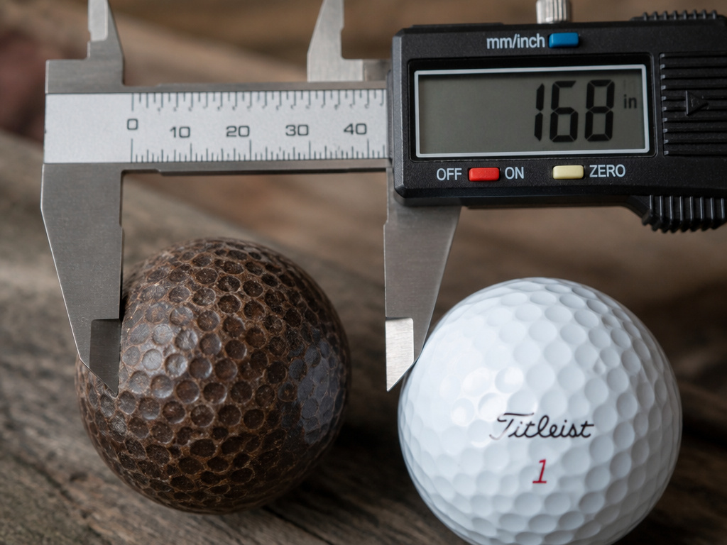 Caliper measuring historical and modern golf balls to show size evolution.